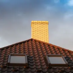 Jasna rura kominowa ceramiczna na ciemnym dachu z dachówkami, pod niebo z chmurami.