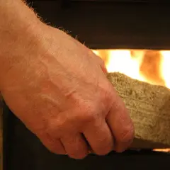 Dłoń wkłada brykiet drzewny liściasty do rozpalonego pieca, gdzie płomienie ogrzewają wnętrze.