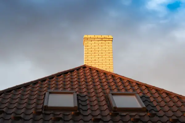 Jasna rura kominowa ceramiczna na ciemnym dachu z dachówkami, pod niebo z chmurami.
